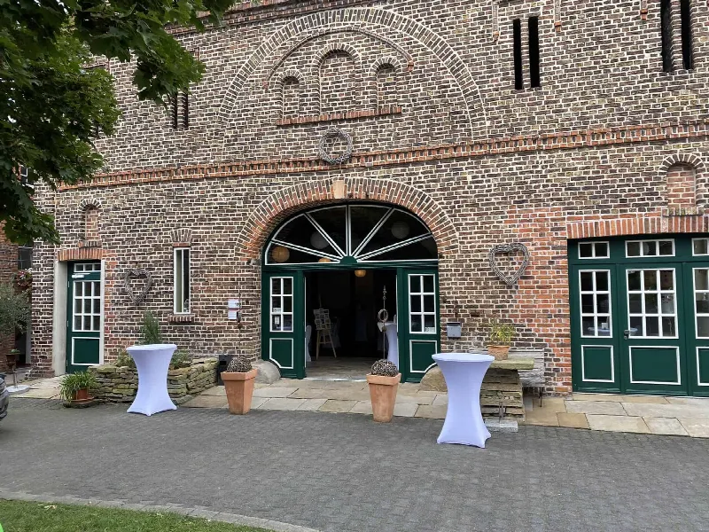 Backsteinfassade mit grünen Türen und Rundbogen, davor zwei Stehtische mit weißen Hussen auf Pflaster