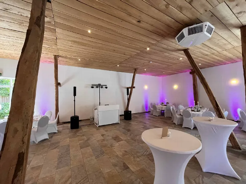 Festscheune mit Holzdecke und Balken, lila Uplights an der Wand, weißes DJ-Pult und Lautsprecher