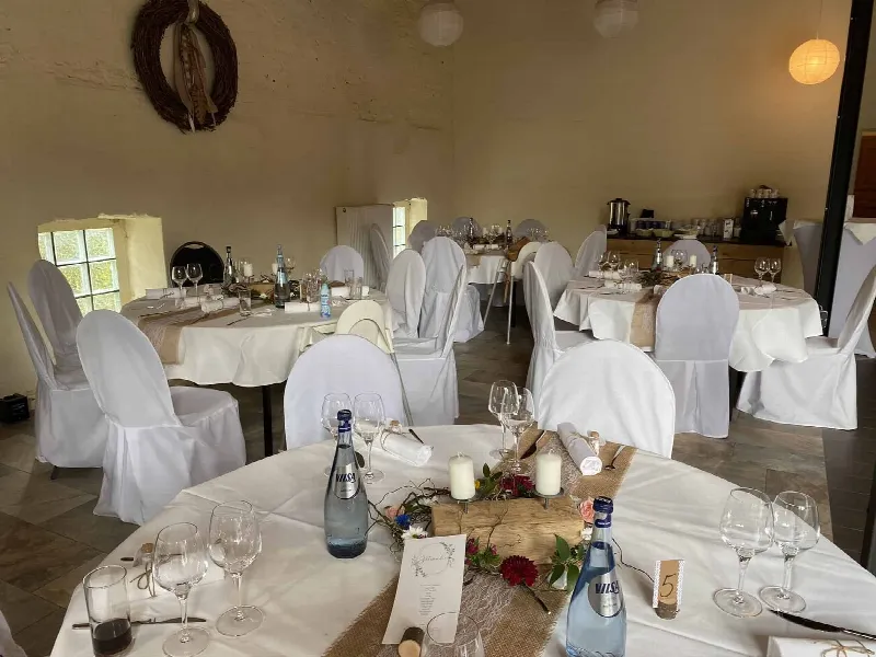 Runde Tische mit Stuhlhussen, Holzdeko mit Blumen und Kerzen, im Hintergrund Kaffeestation