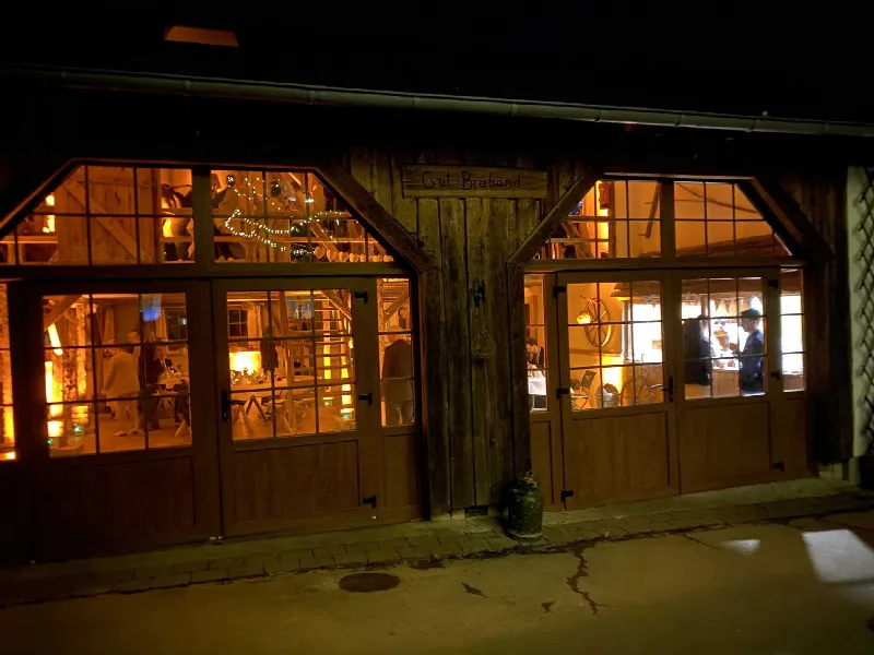 Beleuchtete Scheune mit Holzschild Gut Braband bei Nacht, durch die Fenster sieht man Gäste und Lichterkette