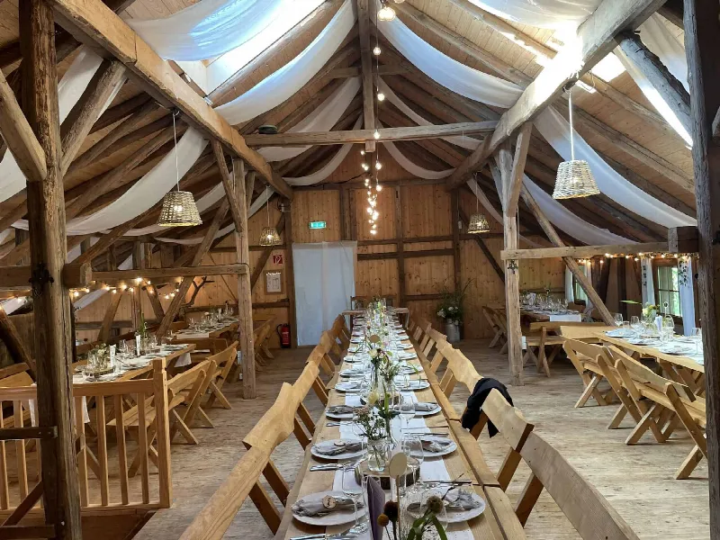 Lange Holztafel mit Bänken in der Scheune, darüber weiße Stoffbahnen, Korblampen und Lichterketten
