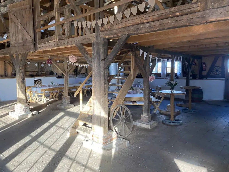 Scheunenhalle mit Holzstützen, Wagenrad und Treppe, im Hintergrund gedeckte Tafeln unter einer Herzgirlande
