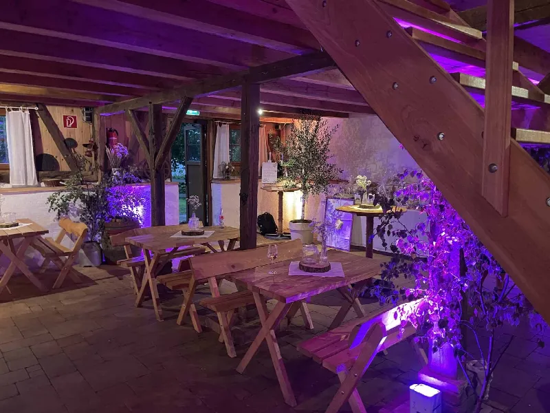 Rustikale Holztische und Bänke unter einer Holztreppe, Pflanzen und Wand werden lila angestrahlt