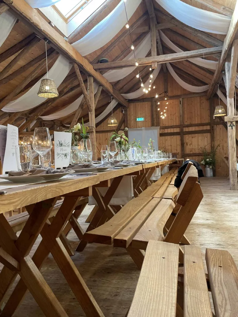 Holztafel und Bänke in der Scheune mit Menükarten, Gläsern, Blumen und Korblampen unter dem Dach