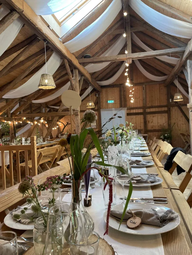 Gedeck mit Namensschild aus Holz, Wildblumen in Vasen und Baumscheibe an der Tafel unter dem Scheunendach