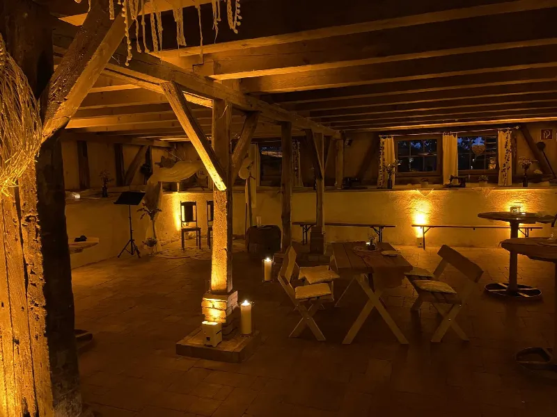 Fachwerkscheune am Abend, Holzpfosten und Wand mit warmem Gelb angestrahlt, Kerzen auf dem Boden und Tischen