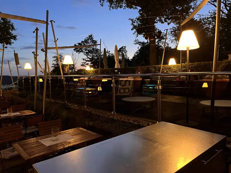 Warm leuchtende Stehlampen und Glasgeländer auf der Terrasse in der Dämmerung, dahinter Hecke und Sonnenschirme