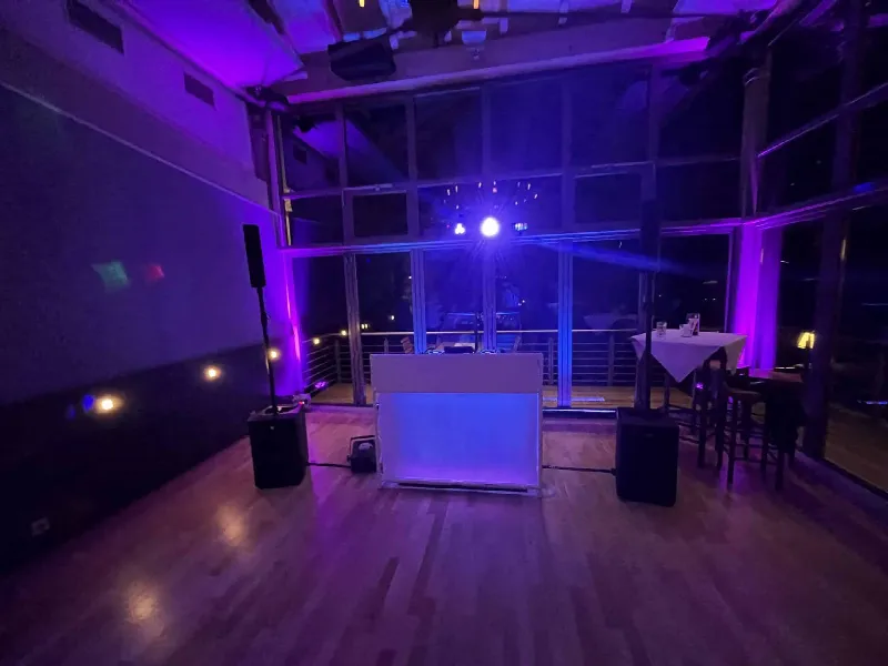 Weißes DJ-Pult mit Lautsprechern auf Parkett im Wintergarten, lila Uplights und Discokugel vor der Glasfront