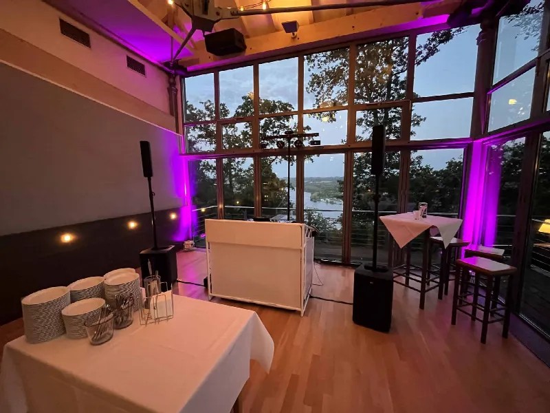 Weißes DJ-Pult und zwei Lautsprecher vor der Glasfront, lila Uplights an den Seiten, Teller auf Tisch links
