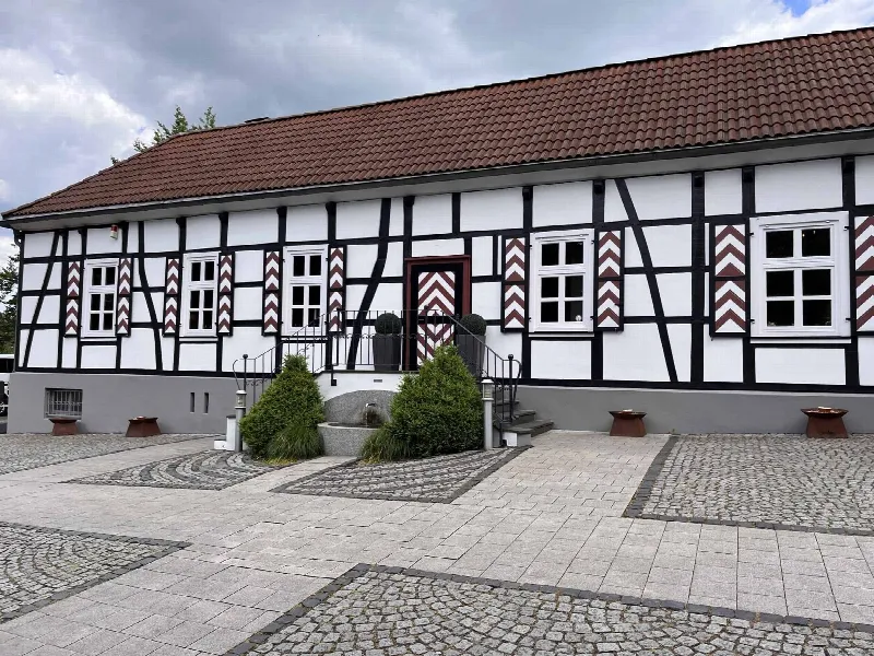 Frontansicht des Fachwerkhauses mit rot-weiß gemusterten Fensterläden, Brunnen und Kopfsteinpflaster