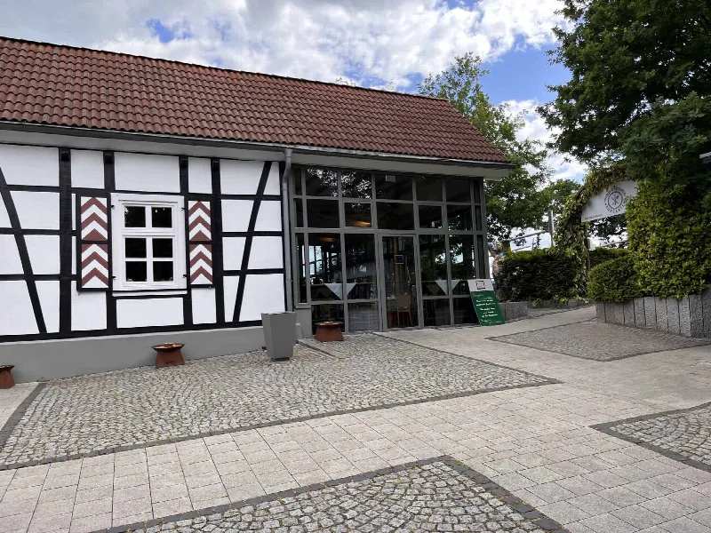 Fachwerkfassade mit Ziegeldach und gläsernem Eingang, rechts Hecke und ein Torbogen mit Schild