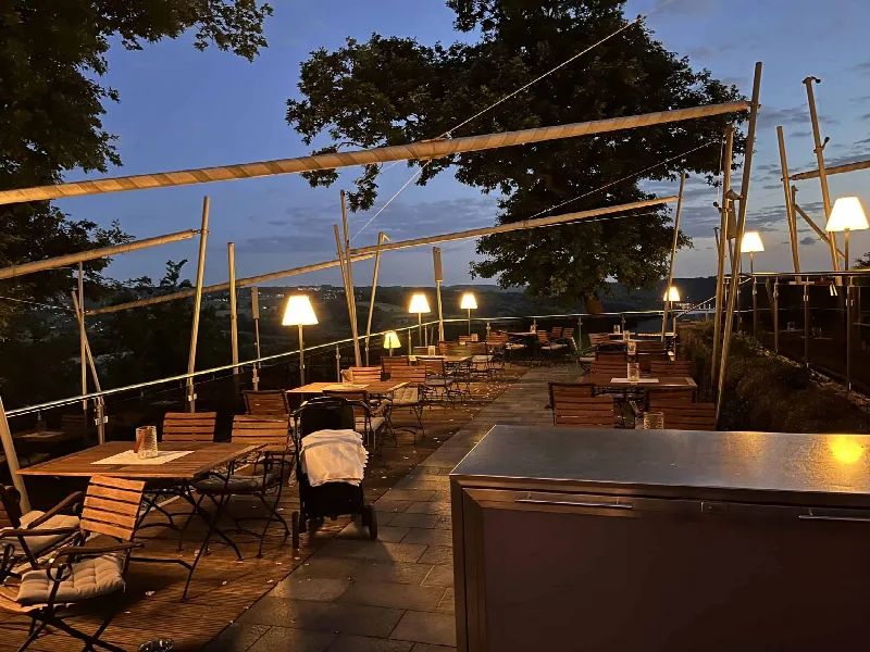 Terrasse mit Holztischen und beleuchteten Stehlampen unter blauem Abendhimmel, vorn ein Edelstahlschrank