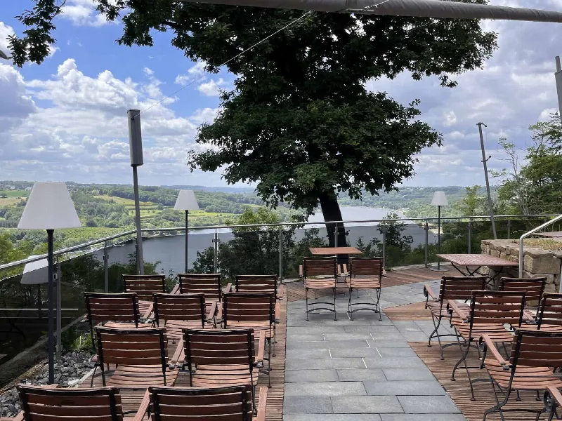 Holzstühle auf der Terrasse unter einer großen Eiche, dahinter Glasgeländer und der Baldeneysee