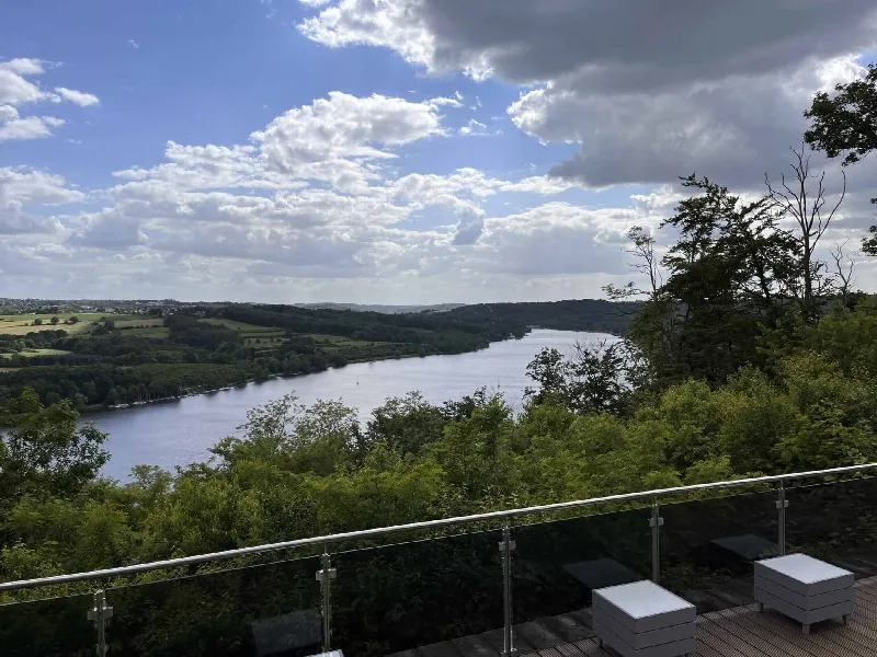 Blick über Glasgeländer und Baumwipfel auf den Baldeneysee, vorn zwei weiße Loungehocker auf Holzdeck