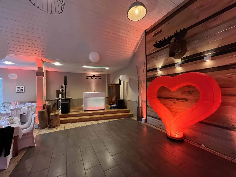 Weißes DJ-Pult mit Lautsprechern und Lichttraverse auf Podest, rechts leuchtet ein rotes Herz an der Holzwand