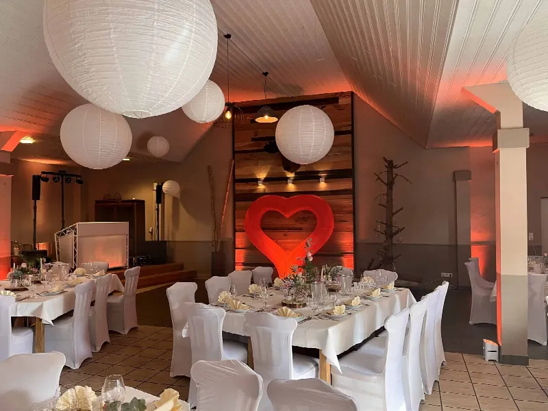 Saal mit lang gedeckter Tafel, großen Papierlampen und leuchtendem Herz vor der Holzwand, im Hintergrund das DJ-Pult