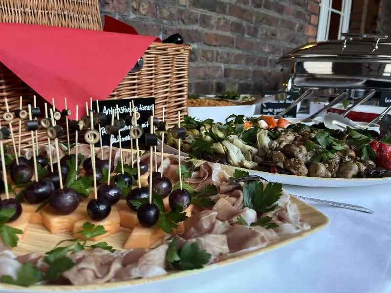 Platte mit Schinken, Melonenspießen mit Oliven und Trauben, daneben marinierte Pilze und Artischocken