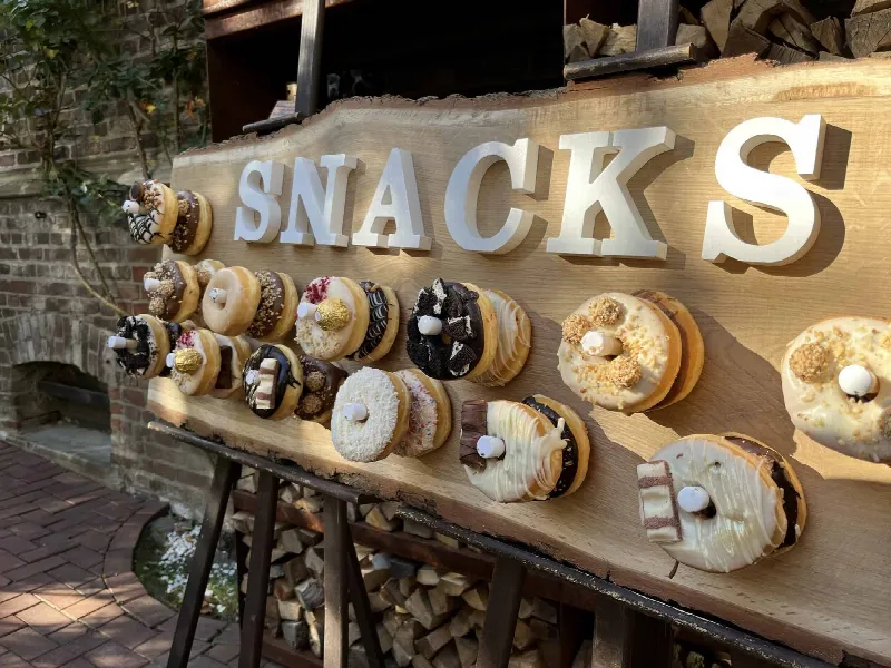 Holzbrett mit weißen Buchstaben „SNACKS“ und bunt dekorierten Donuts auf Stiften vor Backsteinwand