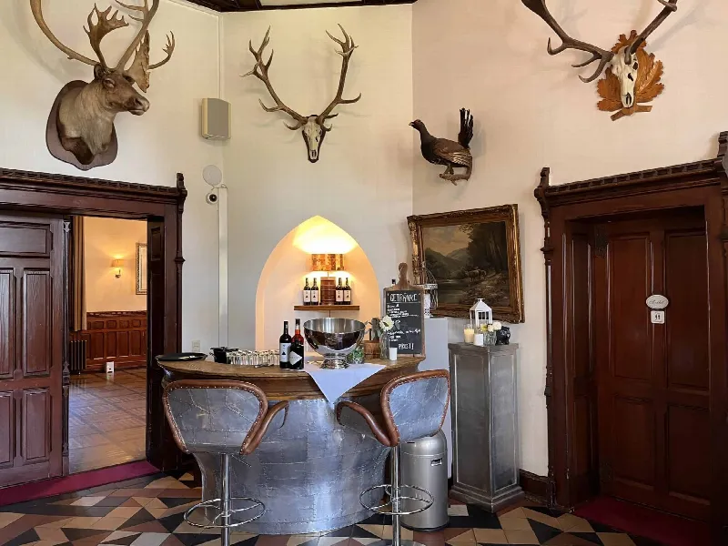 Runde Metallbar mit Barhockern im Foyer, an der Wand Geweihe und Gemälde, daneben Getränketafel