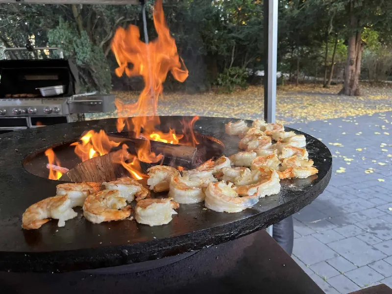 Gambas auf einer Feuerplatte mit Flamme im Freien, im Hintergrund ein Gasgrill und Herbstlaub