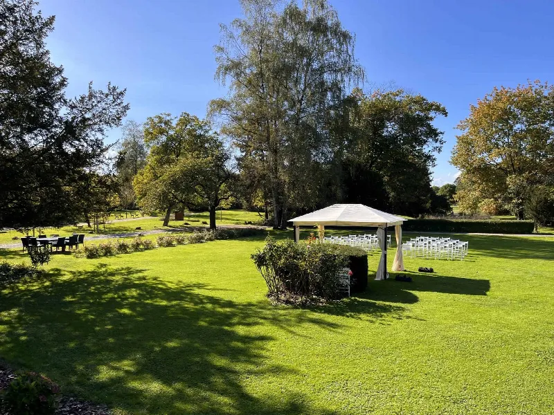 Weißer Pavillon mit weißen Klappstühlen auf gepflegtem Rasen, umgeben von Herbstbäumen unter blauem Himmel
