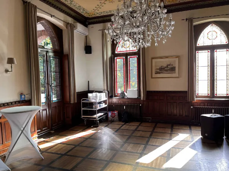 Heller Saal mit Kristallleuchter, bemalter Decke, Buntglasfenstern, Stehtisch und Speisewagen im Sonnenlicht