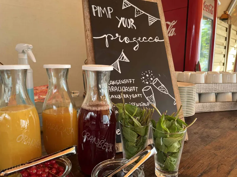 Karaffen mit Orangen-, Maracuja- und Cranberrysaft, Minze und Beeren vor Tafel „Pimp your Prosecco“