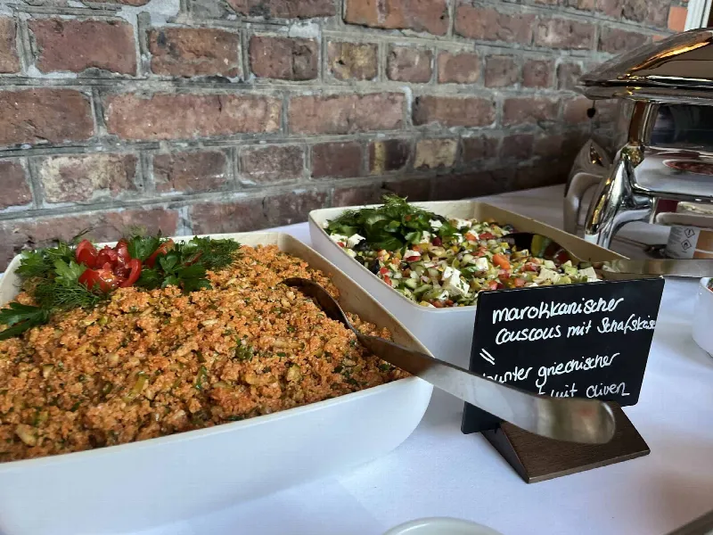 Buffet mit Couscous, griechischem Salat und Kreidetafel, dahinter Chafing Dishes vor Backsteinwand