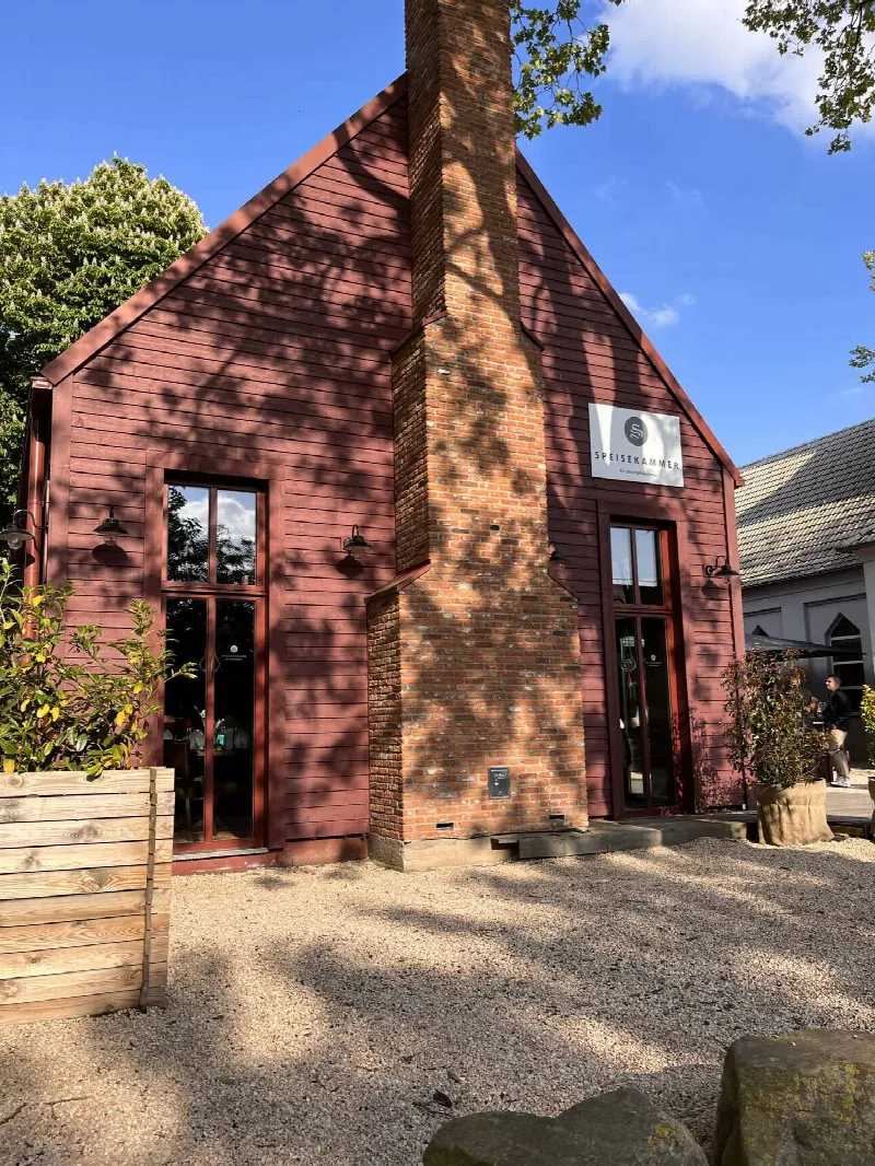 Rotes Holzhaus der Speisekammer mit gemauertem Schornstein, Schriftzug und Kiesplatz bei blauem Himmel