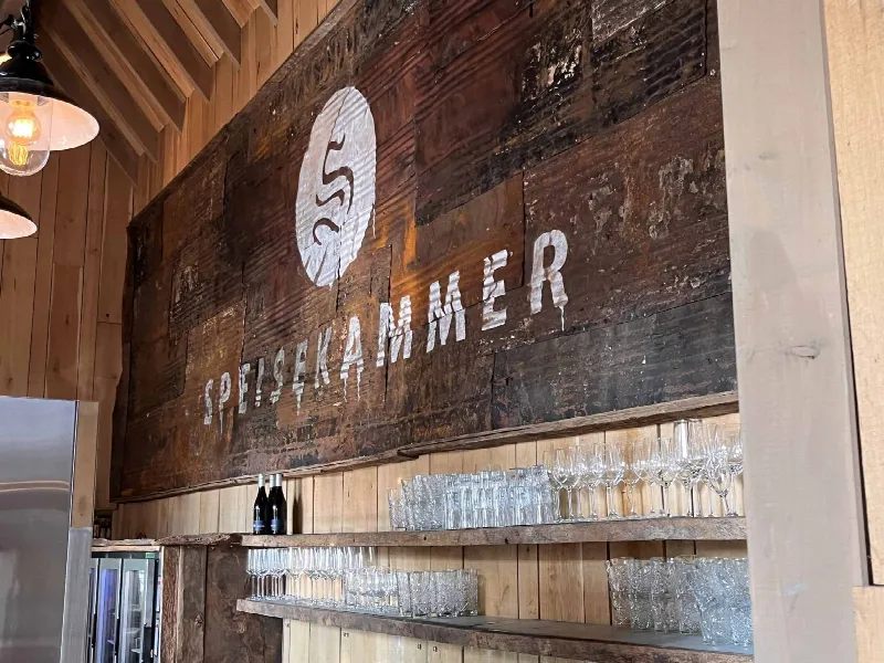 Weißer Schriftzug Speisekammer mit Logo auf dunkler Holzwand über Regalen mit Gläsern und Sektflaschen