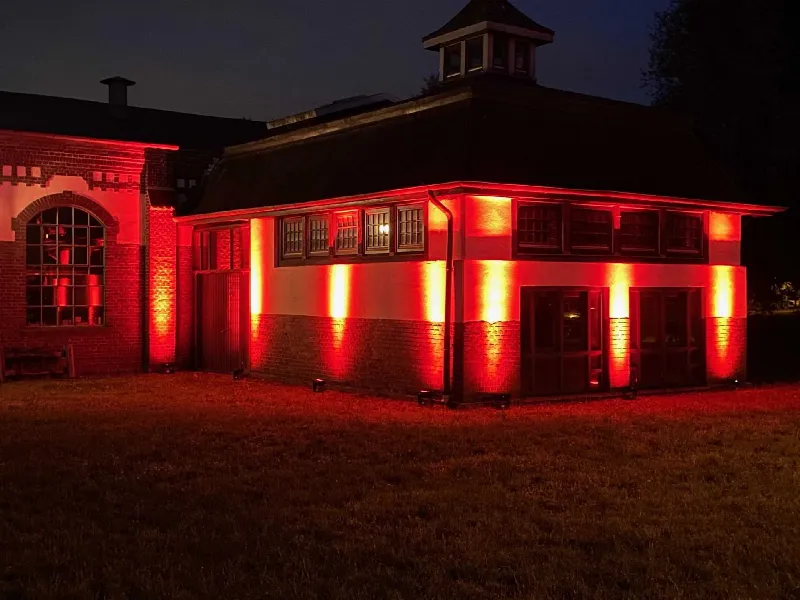 Flaches Backsteingebäude mit Türmchen, mit roten Boden-Strahlern bei Nacht angestrahlt, davor eine Wiese