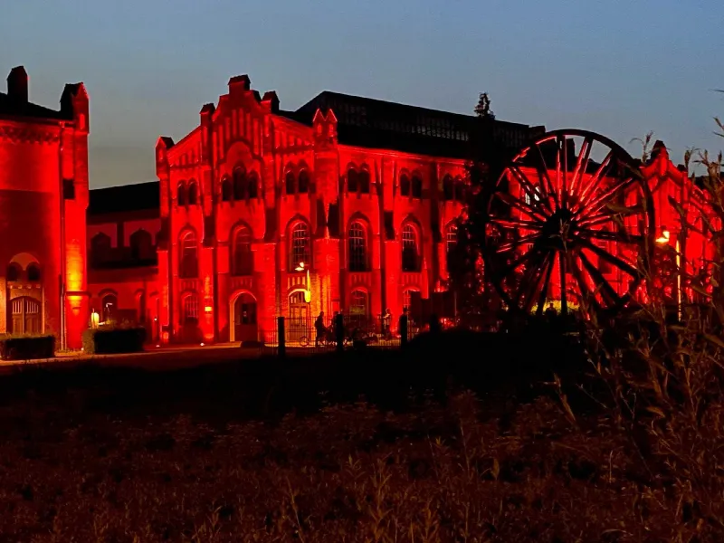 Backsteingebäude der ehemaligen Zeche, rot angestrahlt in der Dämmerung, rechts ein Förderrad